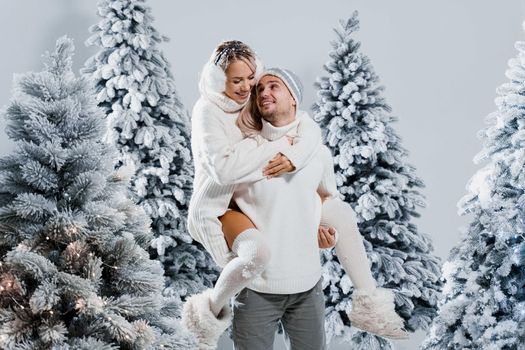 Couple kiss and hug. Man holds girl near christmas trees in winter day. New year celebration. People weared wearing fur headphones, hats, white sweaters.