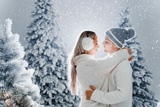 Happy young couple hugs and smiles and snow falls near christmas trees at the eve of new year celebration in winter day. Smiley man and woman weared white pullovers love each other