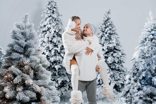 Couple kiss and hug. Man holds girl near christmas trees in winter day. New year celebration. People weared wearing fur headphones, hats, white sweaters.