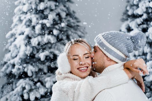 Falling snow and kisses. Happy young couple close-up hugs and kiss near christmas trees at the eve of new year celebration in winter day. Smiley man and woman weared white pullovers love each other