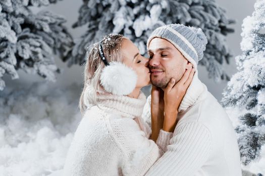 Winter love story with couple weared fur headphones, hats, white sweaters. Happy young couple hugs and kiss near christmas trees at the eve of new year celebration in winter day.