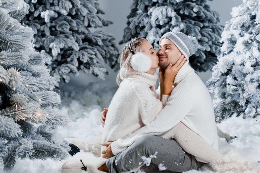 Winter love story with couple weared fur headphones, hats, white sweaters. Happy young couple hugs and kiss near christmas trees at the eve of new year celebration in winter day.