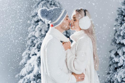 Happy young couple hugs and smiles and snow falls near christmas trees at the eve of new year celebration in winter day. Smiley man and woman weared white pullovers love each other