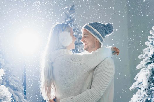 Happy young couple hugs and smiles and snow falls near christmas trees at the eve of new year celebration in winter day. Smiley man and woman weared white pullovers love each other