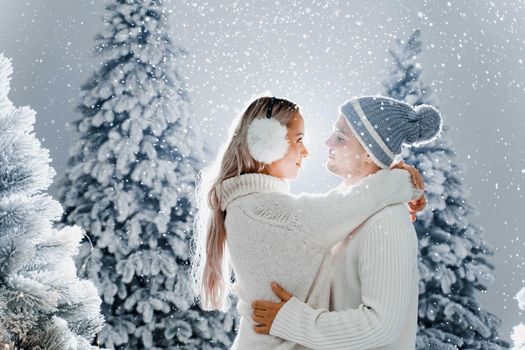 Falling snow and kisses. Happy young couple close-up hugs and kiss near christmas trees at the eve of new year celebration in winter day. Smiley man and woman weared white pullovers love each other