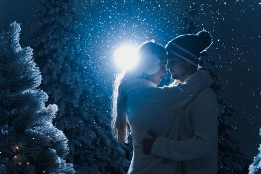 Falling snow and kisses with falling snow and dark blue background. Happy young couple weared white pullovers. hugs and kiss near christmas trees at the eve of new year celebration in winter day