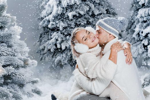 Falling snow and kisses. Happy young couple close-up hugs and kiss near christmas trees at the eve of new year celebration in winter day. Smiley man and woman weared white pullovers love each other