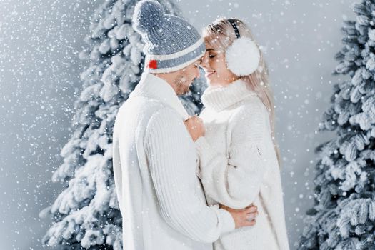 Couple kissing while snow falls near christmass trees. Winter holidays. Love story of young couple weared white pullovers. Happy man and young woman hug each other.