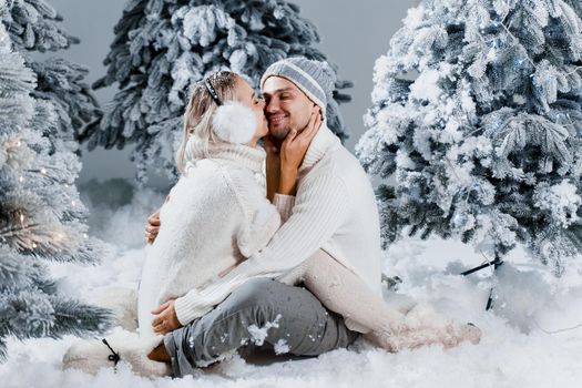 Young woman kissing her lovely man in cheek. Winter holidays. Love story of young couple weared white pullovers. Happy man and young woman hug each other.