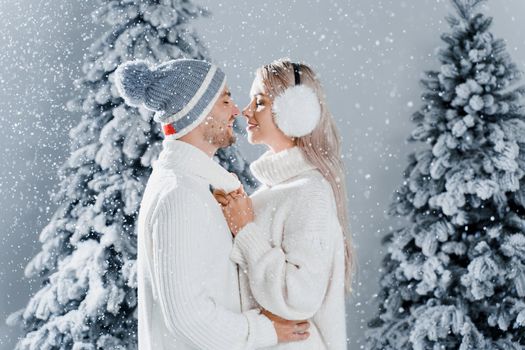 Couple kissing while snow falls near christmass trees. Winter holidays. Love story of young couple weared white pullovers. Happy man and young woman hug each other.