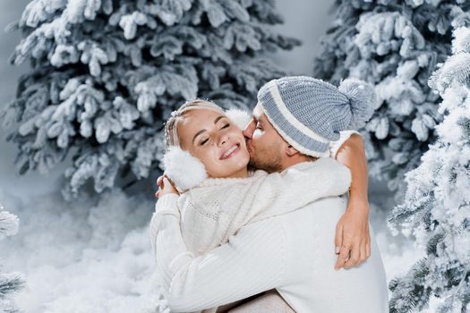 Handsome man is kissing his lovely girl in cheek. Winter holidays. Love story of young couple weared white pullovers. Happy man and young woman hug each other.