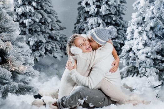 Couple couple laughing and having fun while snow falls near christmass trees. Winter holidays. Love story of young couple weared white pullovers. Happy man and young woman hug each other.