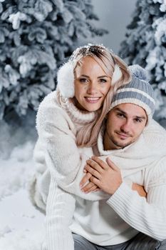 New year love story. Couple kisses and hugs. People weared wearing fur headphones, hats, white sweaters. Happy young couple hugs and kiss near christmas trees in winter day.