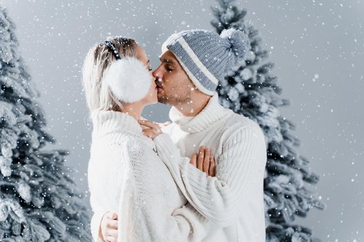 Couple kissing while snow falls near christmass trees. Winter holidays. Love story of young couple weared white pullovers. Happy man and young woman hug each other.