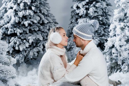 New year love story. Couple kisses and hugs. People weared wearing fur headphones, hats, white sweaters. Happy young couple hugs and kiss near christmas trees in winter day.