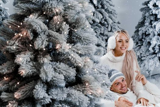 New year love story. Couple kisses and hugs. People weared wearing fur headphones, hats, white sweaters. Happy young couple hugs and kiss near christmas trees in winter day.