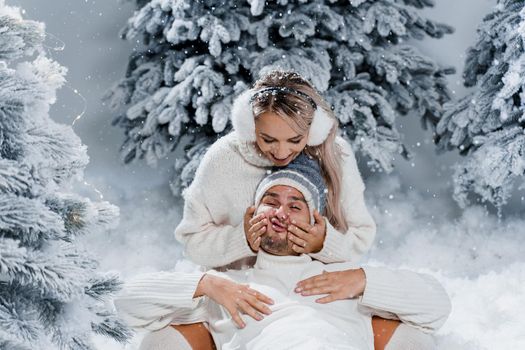 Couple couple laughing and having fun while snow falls near christmass trees. Winter holidays. Love story of young couple weared white pullovers. Happy man and young woman hug each other.