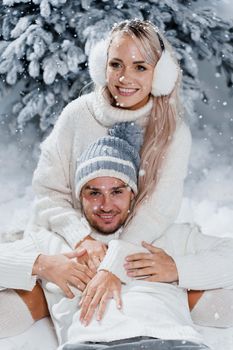 New year love story. Couple kisses and hugs. People weared wearing fur headphones, hats, white sweaters. Happy young couple hugs and kiss near christmas trees in winter day.