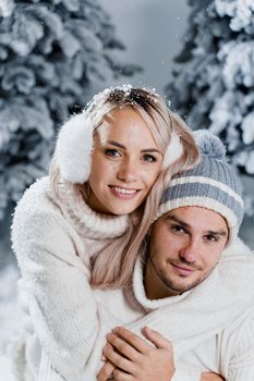 New year love story. Couple kisses and hugs. People weared wearing fur headphones, hats, white sweaters. Happy young couple hugs and kiss near christmas trees in winter day.