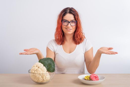 Caucasian woman prefers healthy food. Redhead girl chooses between broccoli and donuts on white background