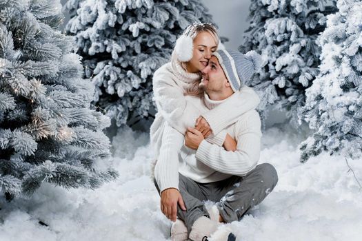 New year love story. Couple kisses and hugs. People weared wearing fur headphones, hats, white sweaters. Happy young couple hugs and kiss near christmas trees in winter day.