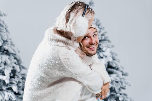 Winter love story with couple weared fur headphones, hats, white sweaters. Happy young couple hugs and kiss near christmas trees at the eve of new year celebration in winter day.