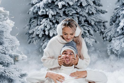 Couple couple laughing and having fun while snow falls near christmass trees. Winter holidays. Love story of young couple weared white pullovers. Happy man and young woman hug each other.