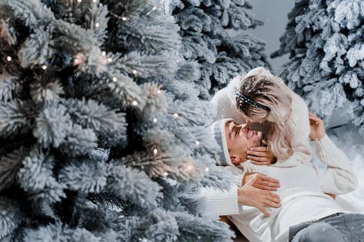 Couple kissing while snow falls near christmass trees. Winter holidays. Love story of young couple weared white pullovers. Happy man and young woman hug each other.