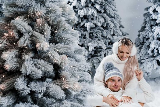 New year love story. Couple kisses and hugs. People weared wearing fur headphones, hats, white sweaters. Happy young couple hugs and kiss near christmas trees in winter day.