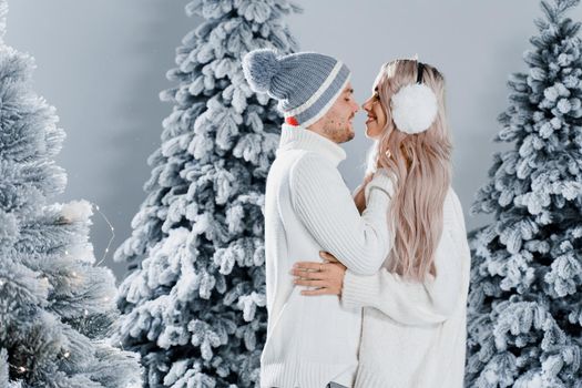 Couple kissing near christmass trees. Winter holidays. Love story of young couple weared white pullovers. Happy man and young woman hug each other.