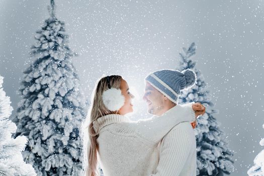 Couple kissing while snow falls near christmass trees. Winter holidays. Love story of young couple weared white pullovers. Happy man and young woman hug each other.