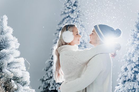 Couple kissing while snow falls near christmass trees. Winter holidays. Love story of young couple weared white pullovers. Happy man and young woman hug each other.