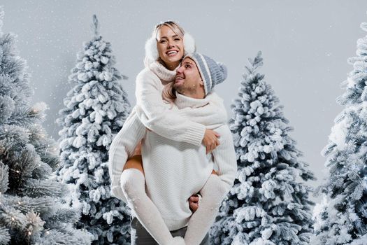 Couple kiss and hug. Man holds girl near christmas trees in winter day. New year celebration. People weared wearing fur headphones, hats, white sweaters.