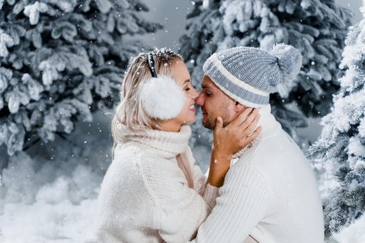 Couple couple laughing and having fun while snow falls near christmass trees. Winter holidays. Love story of young couple weared white pullovers. Happy man and young woman hug each other.