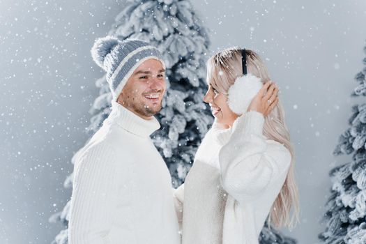 Couple couple laughing and having fun while snow falls near christmass trees. Winter holidays. Love story of young couple weared white pullovers. Happy man and young woman hug each other.