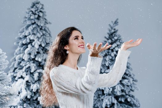 Happy girl smiles and touches falling snow at eve new year. Young woman near big snowy spruces in winter day before christmas celebration