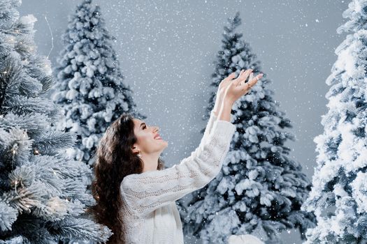 Happy girl smiles and touches falling snow at eve new year. Young woman near big snowy spruces in winter day before christmas celebration