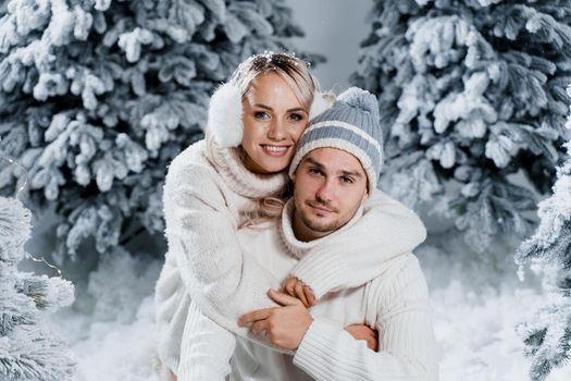 Couple kissing near christmass trees. Winter holidays. Love story of young couple weared white pullovers. Happy man and young woman hug each other.