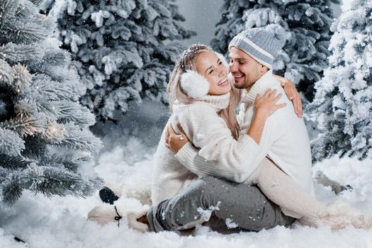 Couple couple laughing and having fun while snow falls near christmass trees. Winter holidays. Love story of young couple weared white pullovers. Happy man and young woman hug each other.