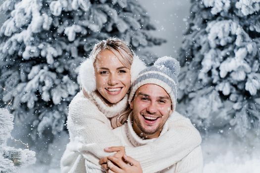Couple couple laughing and having fun while snow falls near christmass trees. Winter holidays. Love story of young couple weared white pullovers. Happy man and young woman hug each other.