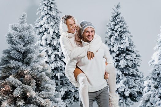 Couple couple laughing and having fun while snow falls near christmass trees. Winter holidays. Love story of young couple weared white pullovers. Happy man and young woman hug each other.