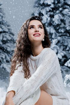 Attractive girl in a warm white sweater and white socks near snowy trees before the new year. Christmas holiday.