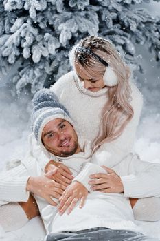 Couple couple laughing and having fun while snow falls near christmass trees. Winter holidays. Love story of young couple weared white pullovers. Happy man and young woman hug each other.