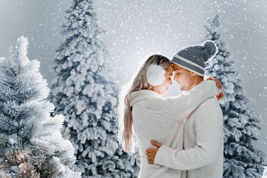 Couple kissing while snow falls near christmass trees. Winter holidays. Love story of young couple weared white pullovers. Happy man and young woman hug each other.