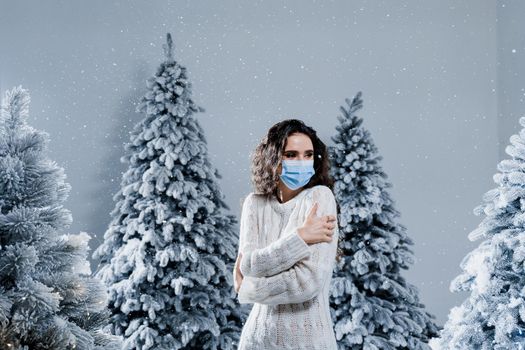 New year celebration at covid-19 coronavirus quarantine period. Happy girl in medical mask with falling snow stays at home. Social distance. Winter holidays in snowy day