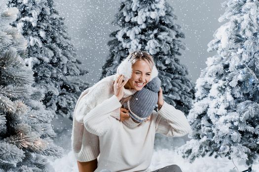 Couple couple laughing and having fun while snow falls near christmass trees. Winter holidays. Love story of young couple weared white pullovers. Happy man and young woman hug each other.