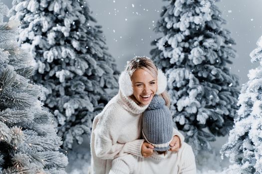 Couple couple laughing and having fun while snow falls near christmass trees. Winter holidays. Love story of young couple weared white pullovers. Happy man and young woman hug each other.