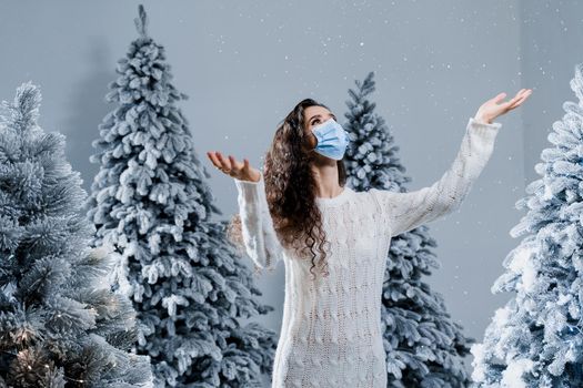 New year celebration at covid-19 coronavirus quarantine period. Happy girl in medical mask with falling snow stays at home. Social distance. Winter holidays in snowy day