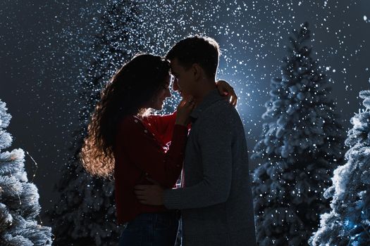 Falling snow and kisses with falling snow on dark blue background. Happy young couple hug and kiss near christmas trees at the eve of new year celebration in winter day