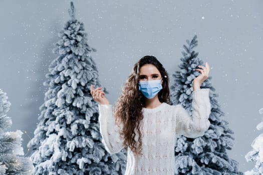 New year celebration at covid-19 coronavirus quarantine period. Happy girl in medical mask with falling snow stays at home. Social distance. Winter holidays in snowy day
